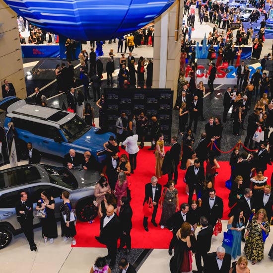 2026 First Look for Charity Fundraiser Raises Over $2 Million for Local Charities; A bustling gala event with people in formal attire on a red carpet surrounded by cars and a large blue structure overhead, exuding elegance and excitement.