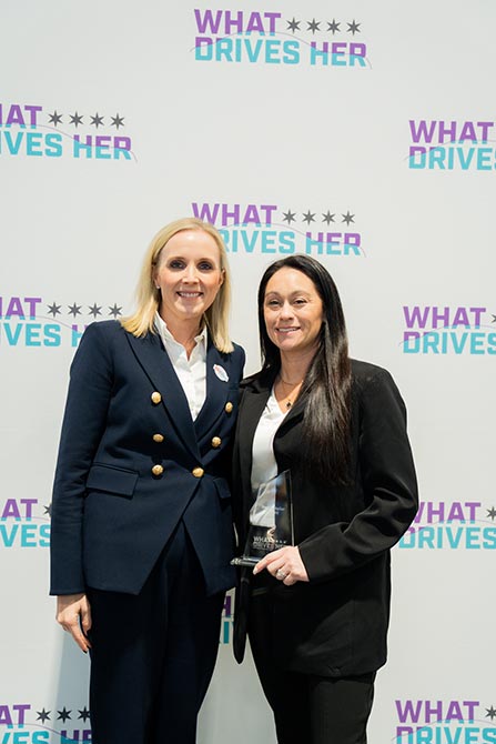Two women standing in front of a "What Drives Her" backdrop, smiling warmly. They are dressed in business attire, conveying empowerment and unity.