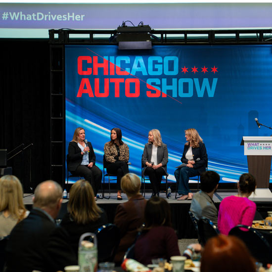 2026 What Drives Her Award Winners; Four women sit on stage in discussion at the Chicago Auto Show, with a banner reading "#WhatDrivesHer." Audience members watch attentively.
