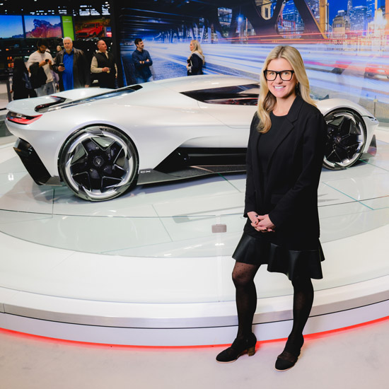 Letter From the President: A Strong Show and a Busy Season Ahead; A woman in black stands smiling in front of a sleek, silver concept car on display. The background is lively with people and futuristic city images.