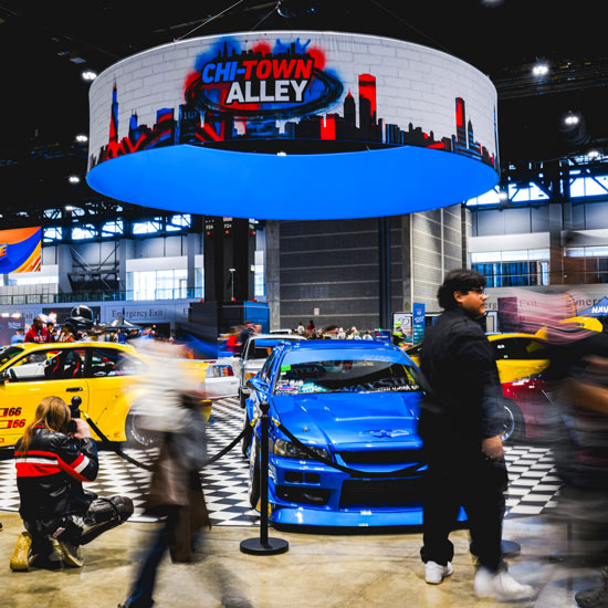 Chi-Town Alley Debuts at the 2026 Chicago Auto Show; Car show scene titled "Chi-Town Alley" with blue and yellow custom cars on checkered floor. Enthusiasts and photographers bustle around excitedly.