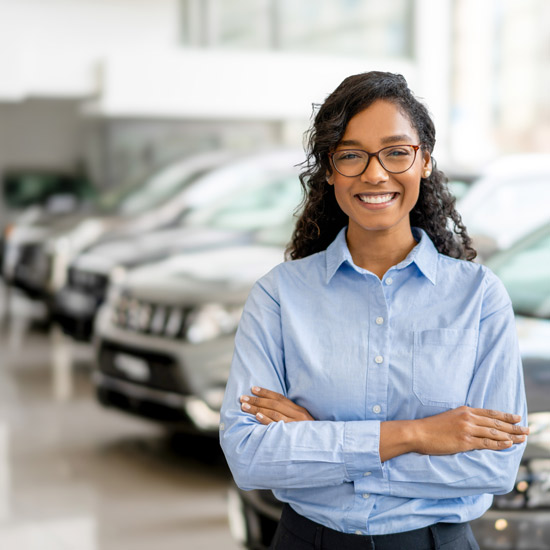CATA Drive for Excellence Awards; Smiling woman in glasses and a blue shirt stands with arms crossed in a car dealership. Several cars are visible in the blurred background.